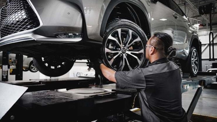 Lexus-certified technician adjusting front driver-side wheel alignment on a lifted silver Lexus SUV in a service bay